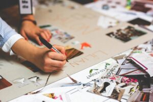 Main féminine écrivant sur un collage entouré de découpes colorées et d’images, dans un atelier créatif.
