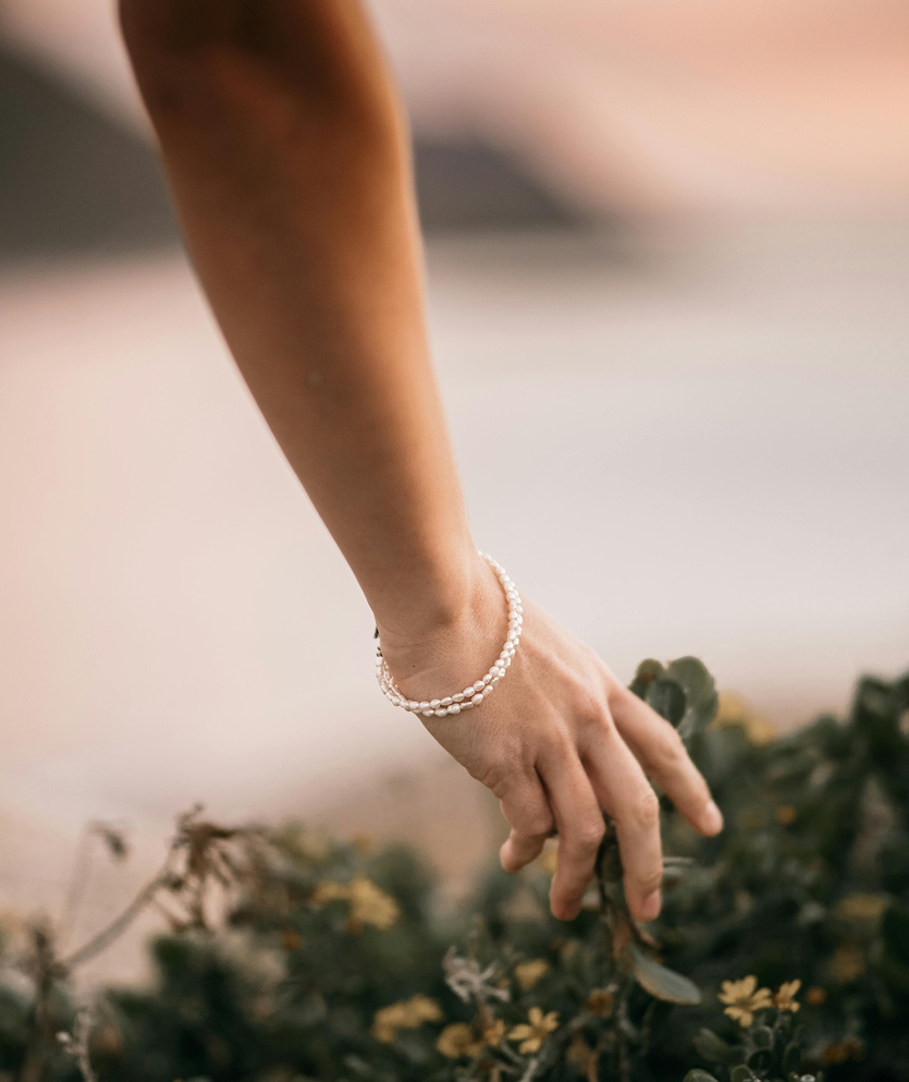 Main féminine portant un bracelet de perles, effleurant des plantes vertes et fleuries, symbolisant la connexion avec la nature.