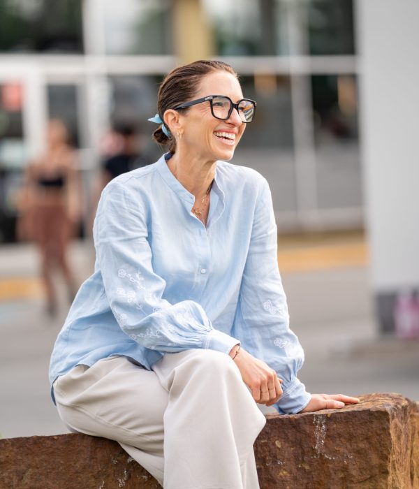 Caroline Blouin – Fondatrice et guide de la retraite bien-être Caroline Blouin, fondatrice de retraites bien-être, souriante et assise à l’extérieur, portant une chemise bleue et des lunettes noires.