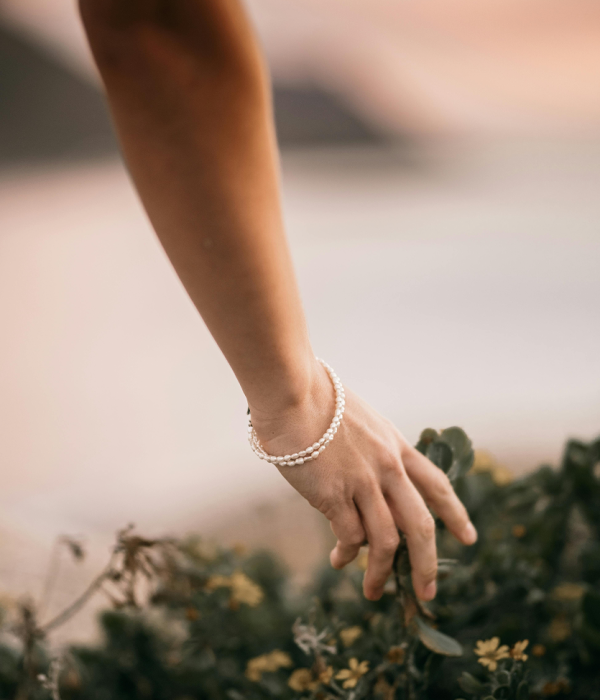 Main délicate effleurant la nature Main féminine portant un bracelet de perles, effleurant des plantes vertes et fleuries, symbolisant la connexion avec la nature.